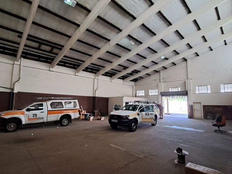 Spacious industrial warehouse or garage area featuring high ceilings with exposed steel trusses, concrete flooring, and large roller shutter access.