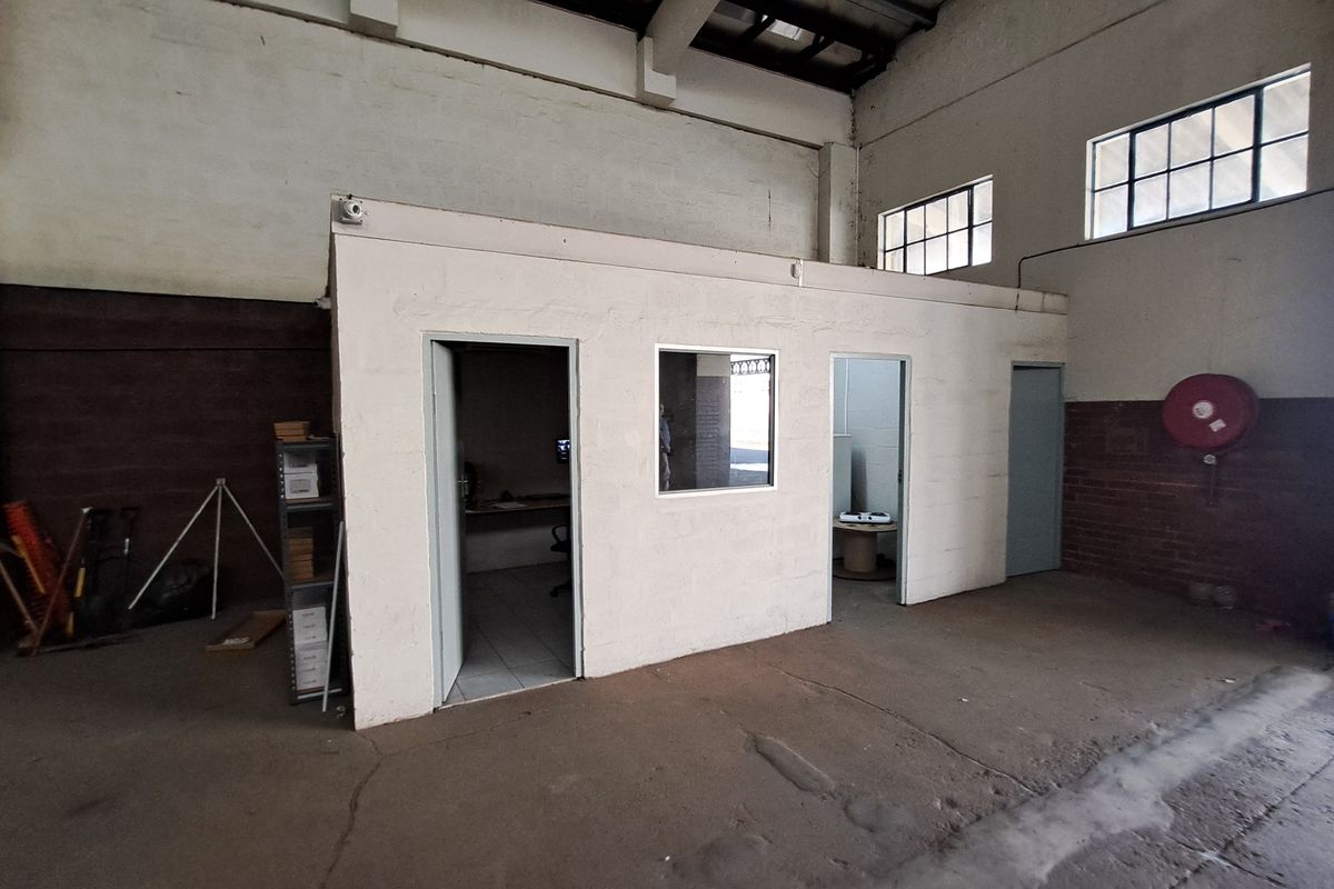 Industrial warehouse space featuring a partitioned office block, exposed steel roof trusses, and concrete flooring, suitable for storage or workshop use.