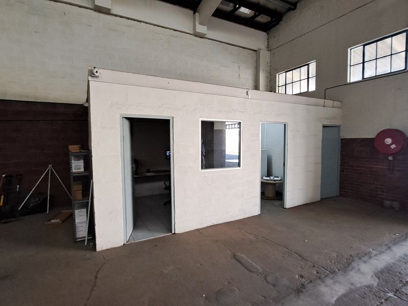 Industrial warehouse space featuring a partitioned office block, exposed steel roof trusses, and concrete flooring, suitable for storage or workshop use.