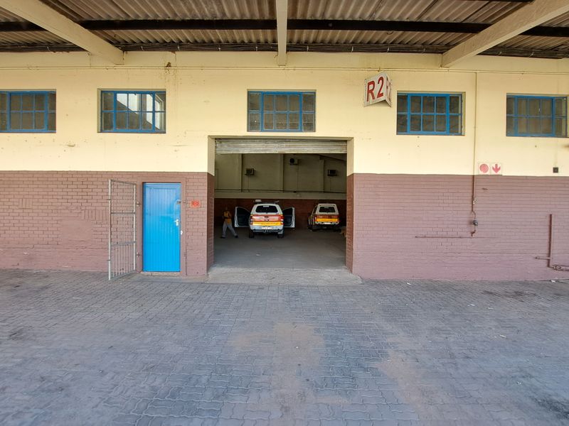 Industrial warehouse exterior featuring a large roller shutter entrance, paved forecourt, and brick facade with steel-framed windows.