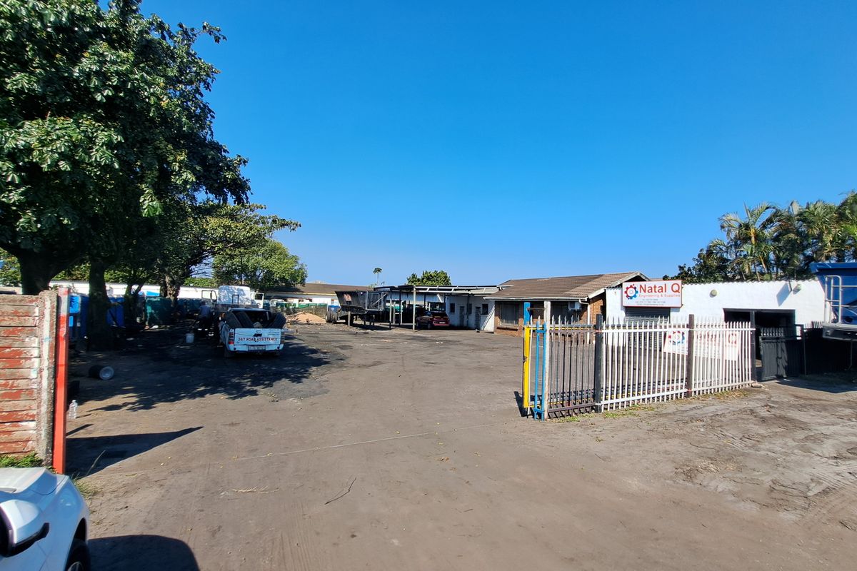 Industrial property exterior featuring a large asphalt yard, metal security fencing, and a functional workshop building with covered loading space.