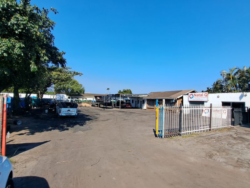 Industrial property exterior featuring a large asphalt yard, metal security fencing, and a functional workshop building with covered loading space.
