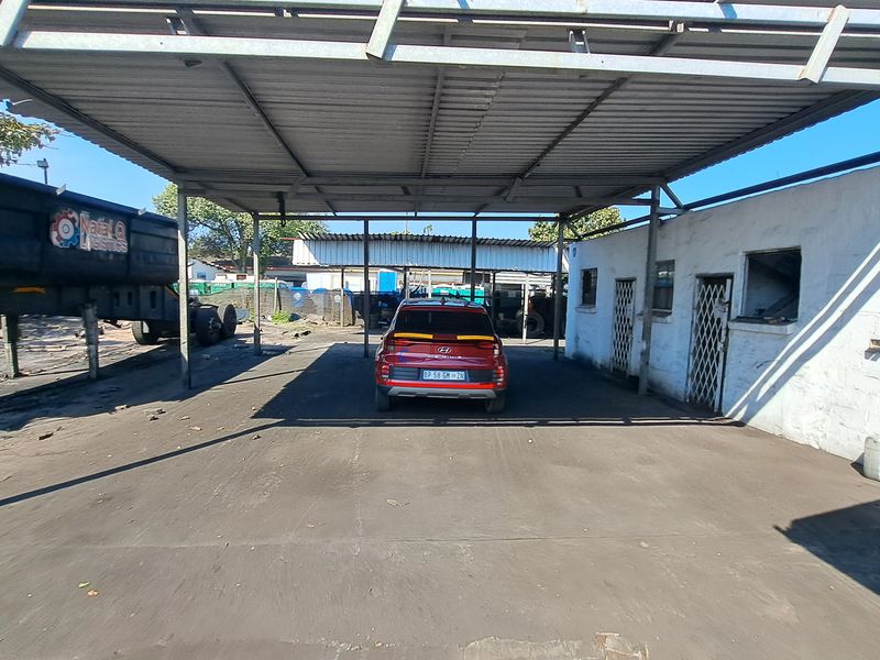 Industrial outdoor covered parking or loading area featuring a metal roof structure, concrete flooring, and adjacent masonry building with security gates.