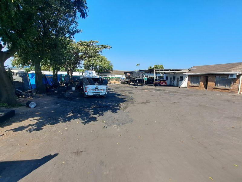 Industrial property exterior featuring a large asphalt yard, covered parking area, and brick building facade with roller shutter access.