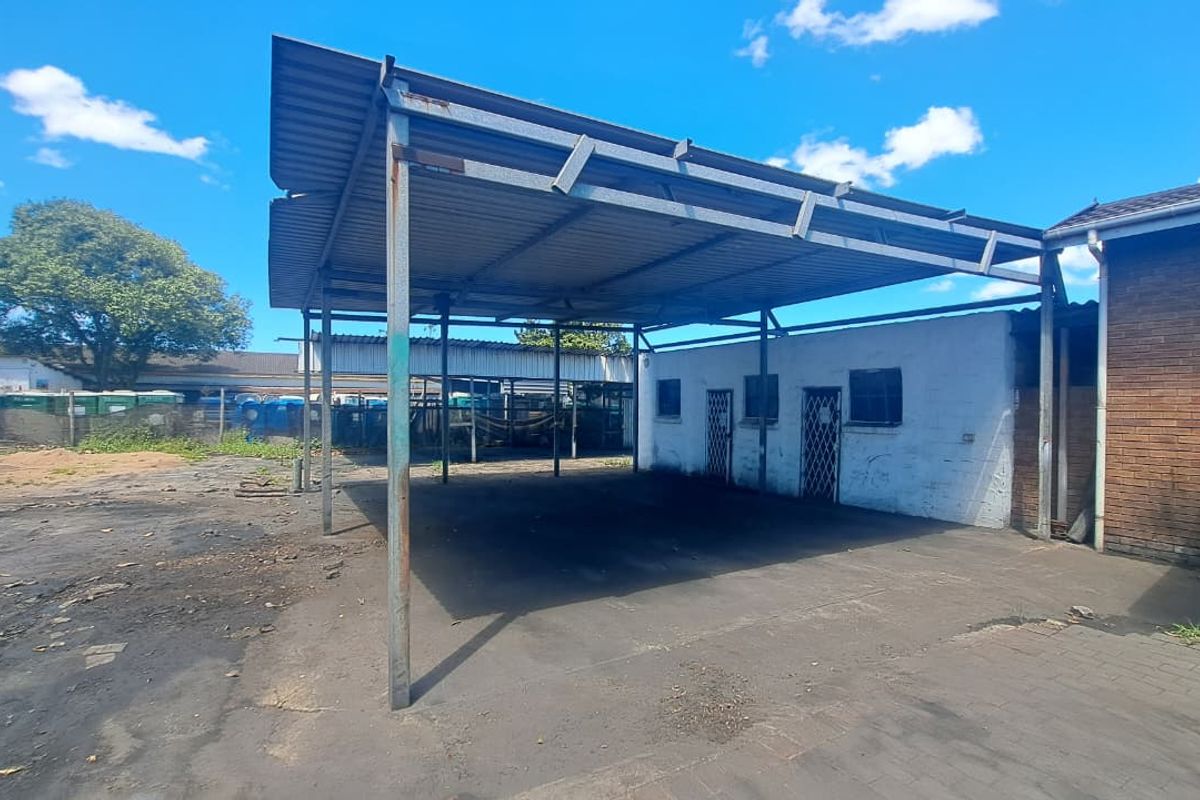 Industrial exterior area featuring a large metal carport structure, concrete paving, and access to adjacent brick and masonry building units.