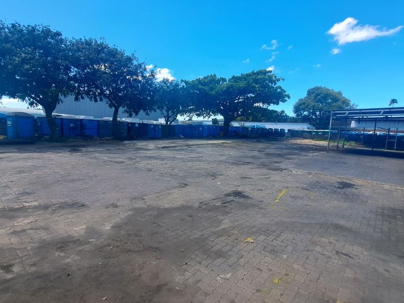 Spacious industrial paved yard area featuring a covered parking structure and mature trees, suitable for storage or vehicle access.