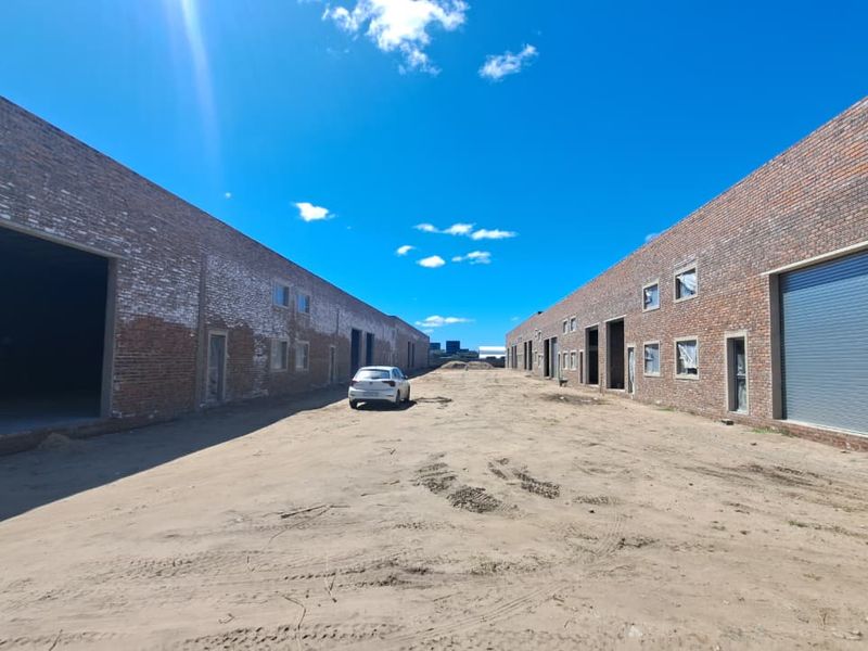 Exterior view of an industrial warehouse complex featuring exposed brick facades, multiple roller shutter doors, and an unpaved yard area.