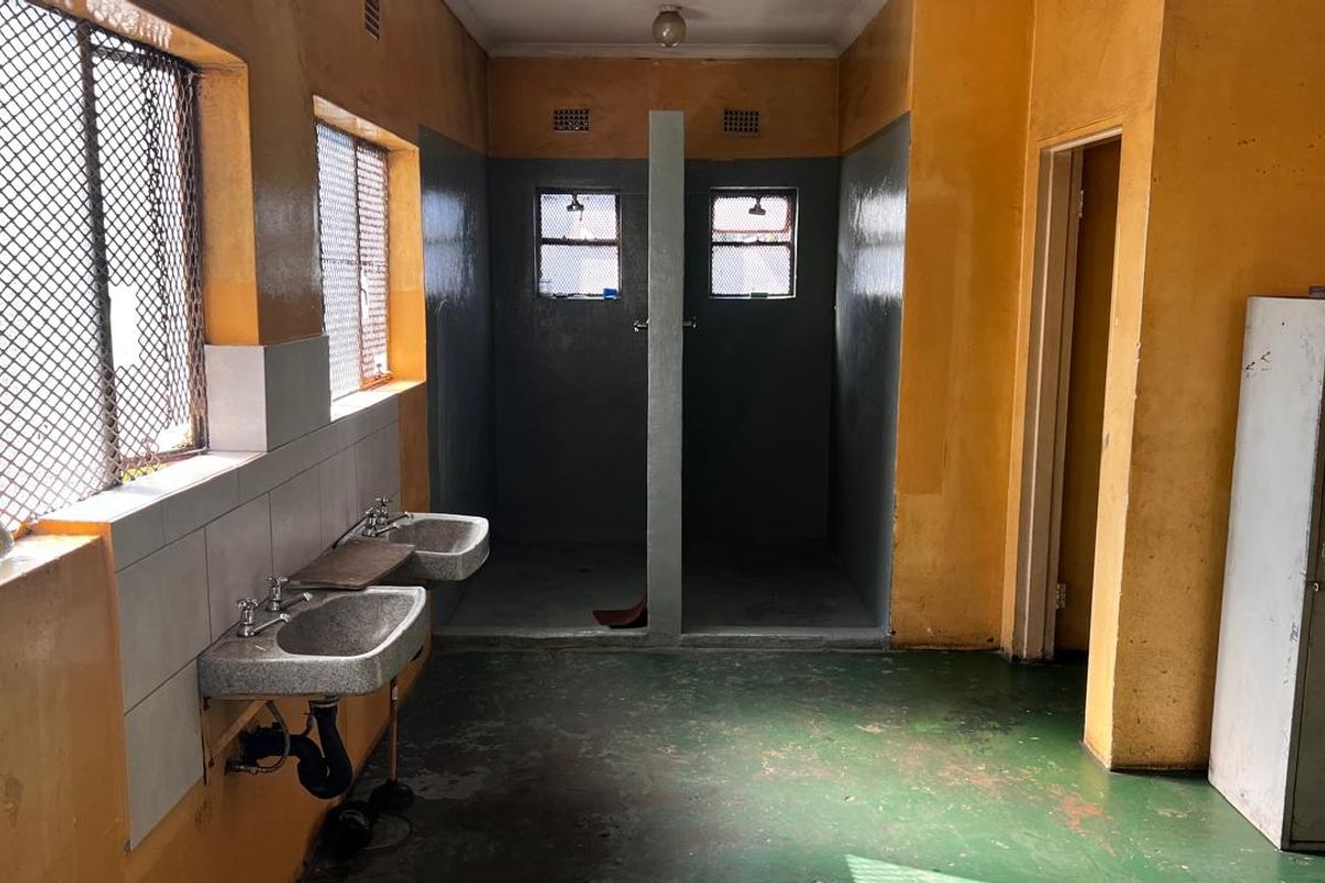 Industrial-style communal bathroom featuring wall-mounted basins, dual shower cubicles, and security-grille windows, requiring significant refurbishment of wall and floor finishes.
