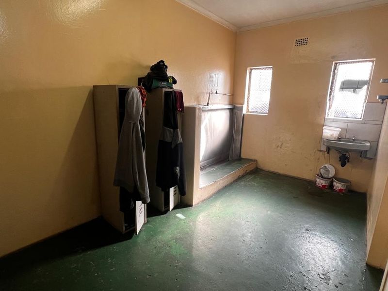 Industrial utility or change room featuring metal lockers, a stainless steel trough urinal, and a wall-mounted basin with security-barred windows.