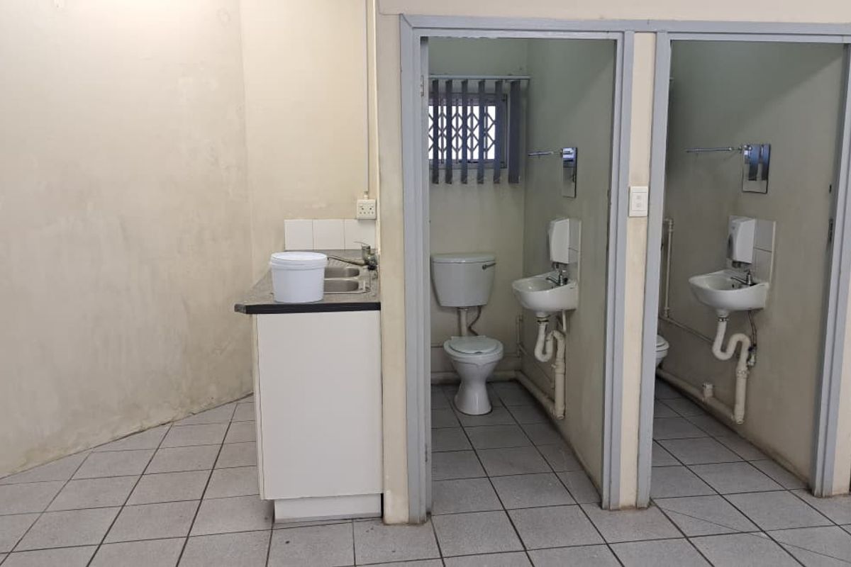 A functional commercial bathroom area featuring two toilet cubicles, wall-mounted basins, and a small kitchenette sink unit with tiled flooring.