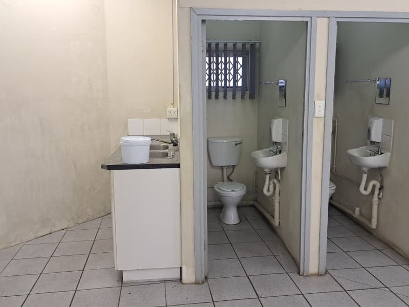 A functional commercial bathroom area featuring two toilet cubicles, wall-mounted basins, and a small kitchenette sink unit with tiled flooring.