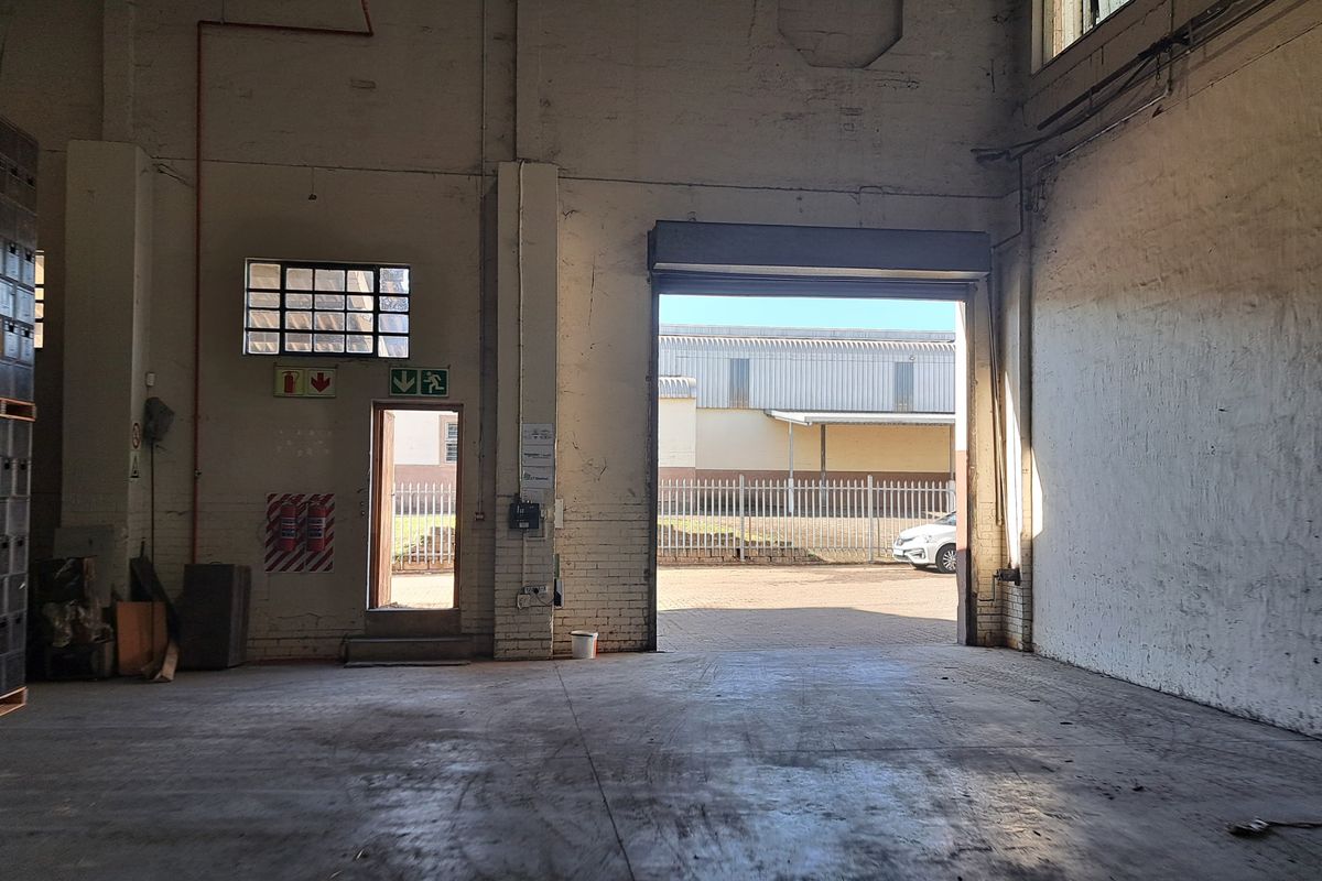 Spacious industrial warehouse unit featuring high ceilings, concrete flooring, a large roller shutter door, and dedicated emergency exit access.