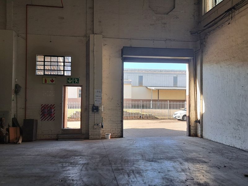 Spacious industrial warehouse unit featuring high ceilings, concrete flooring, a large roller shutter door, and dedicated emergency exit access.