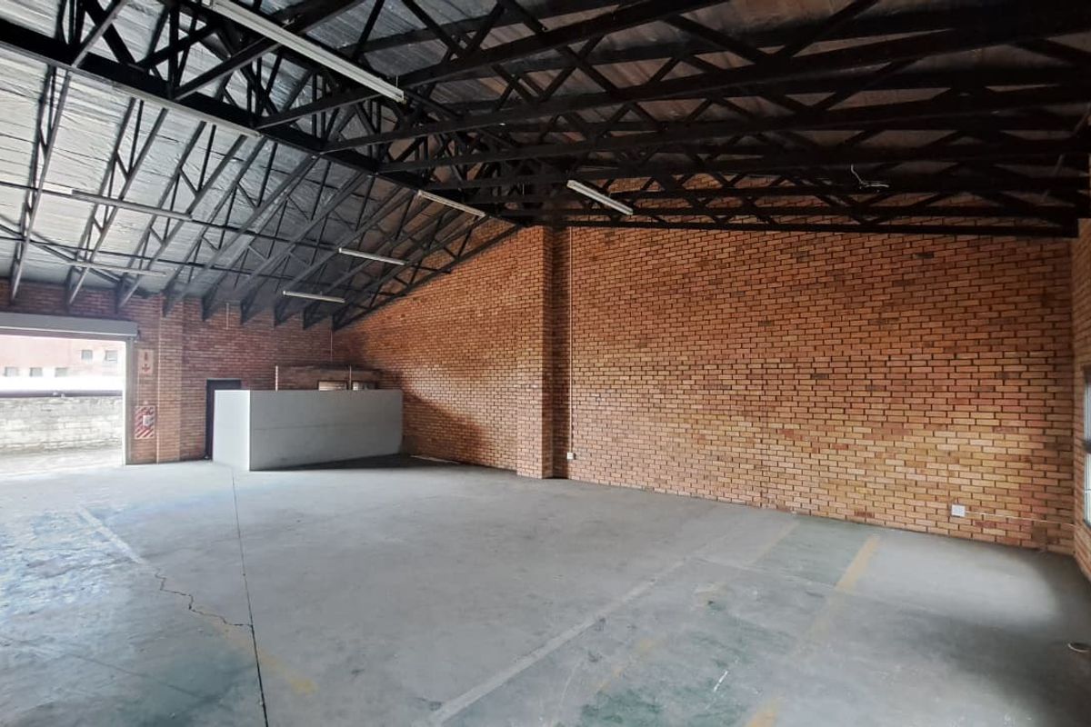 Spacious industrial utility area featuring exposed brick walls, high steel truss ceilings, concrete flooring, and a large roller shutter door for easy access.