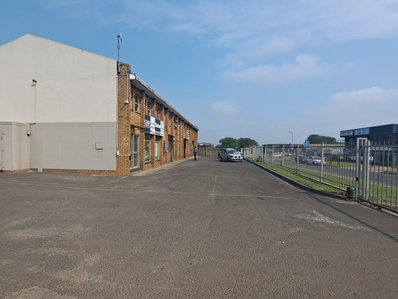 Exterior view of an industrial property featuring a large asphalt parking area, brick facade, and secure perimeter fencing.