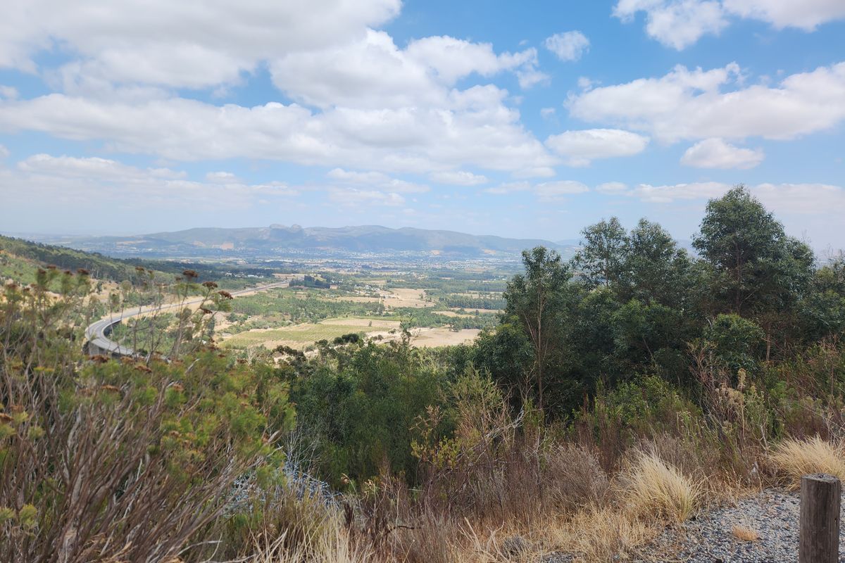 A scenic elevated landscape view overlooking a rural valley, rolling hills, and distant mountains under a bright blue sky.