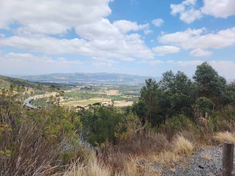 A scenic elevated landscape view overlooking a rural valley, rolling hills, and distant mountains under a bright blue sky.