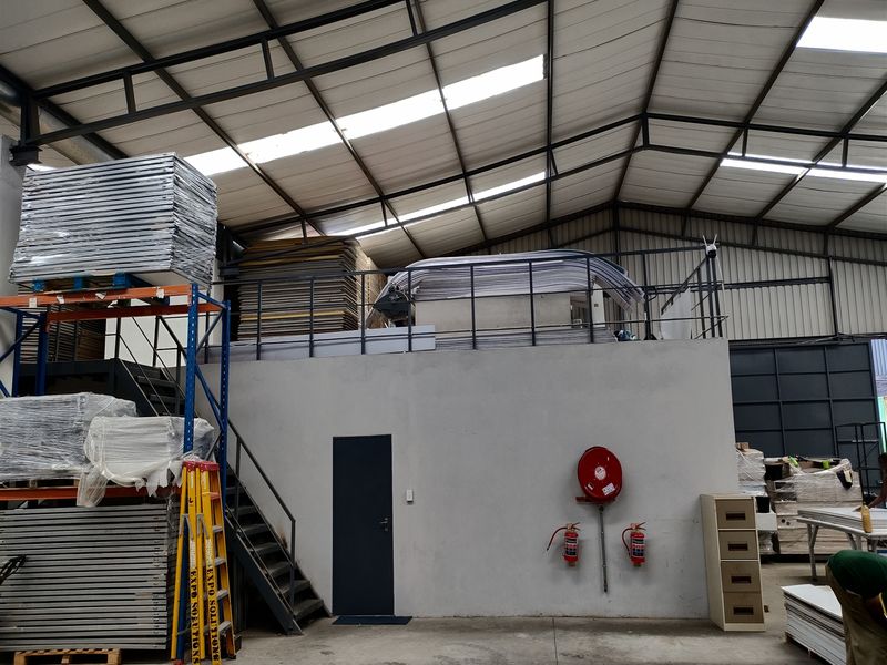 Industrial warehouse space featuring a mezzanine level, steel staircase, exposed roof trusses with skylights, and integrated fire safety equipment.