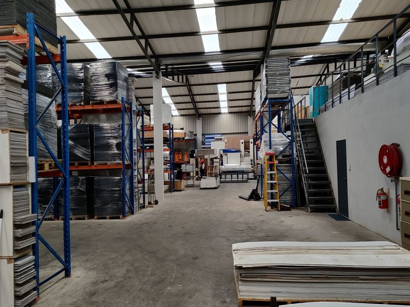 Spacious industrial warehouse featuring high ceilings with integrated skylights, heavy-duty steel racking systems, a mezzanine level with staircase, and durable concrete flooring.