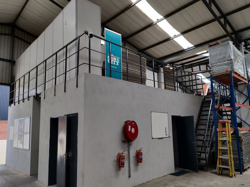 Industrial warehouse interior featuring a mezzanine level with metal staircase, fire safety equipment, and high-clearance roof with integrated skylights.