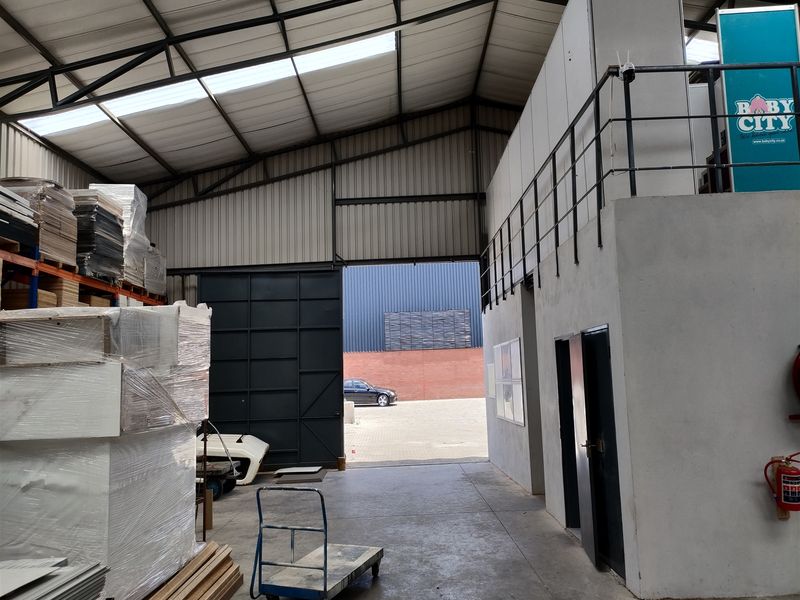 Industrial warehouse space featuring a high-pitched metal roof, skylights, a mezzanine level with railing, and a large sliding industrial access door.