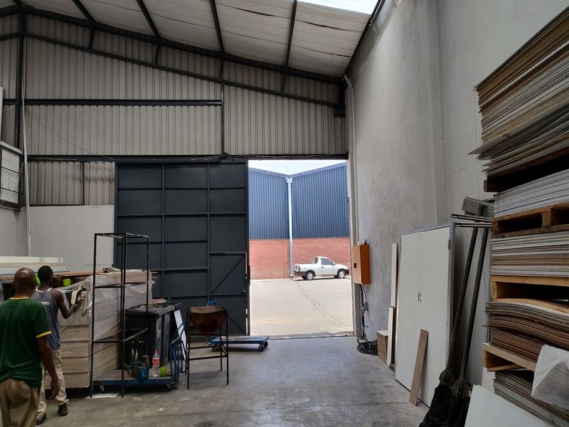 Industrial warehouse space featuring high ceilings with translucent roof panels, concrete flooring, and a large sliding access door for loading.