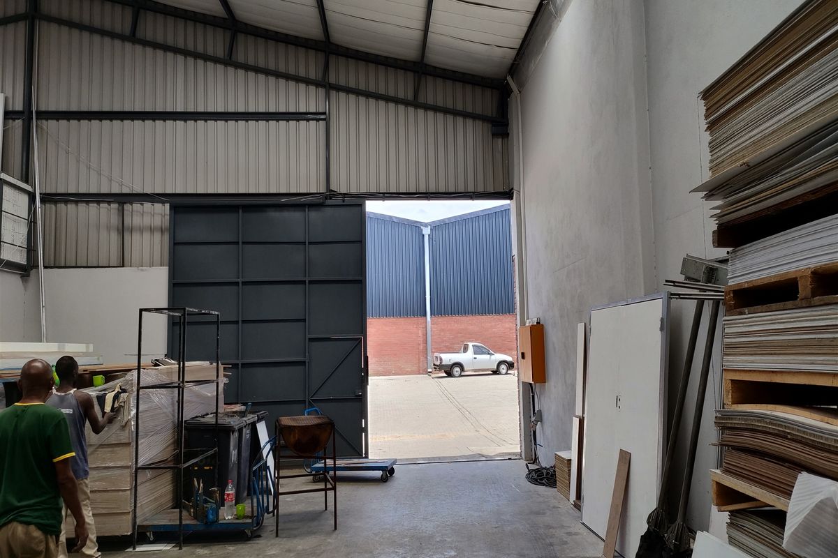 Industrial warehouse space featuring high ceilings with translucent roof panels, concrete flooring, and a large sliding access door for loading.