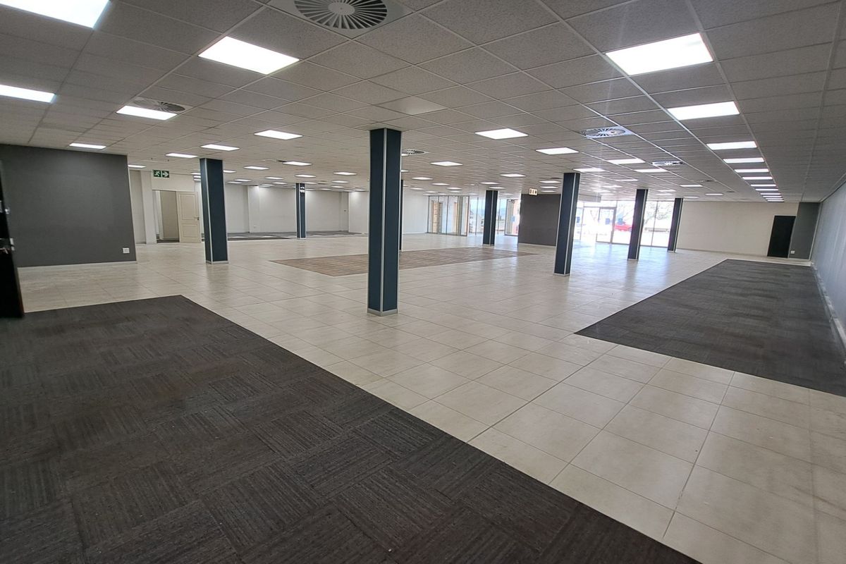 A large, open-plan office space featuring a suspended ceiling, recessed lighting, structural columns, and a combination of tiled and carpeted flooring.
