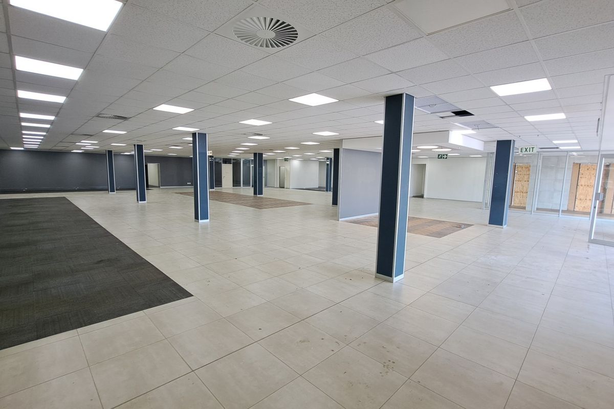 A large, open-plan office space featuring a suspended ceiling with recessed lighting, a mix of tiled and carpeted flooring, and structural support columns.
