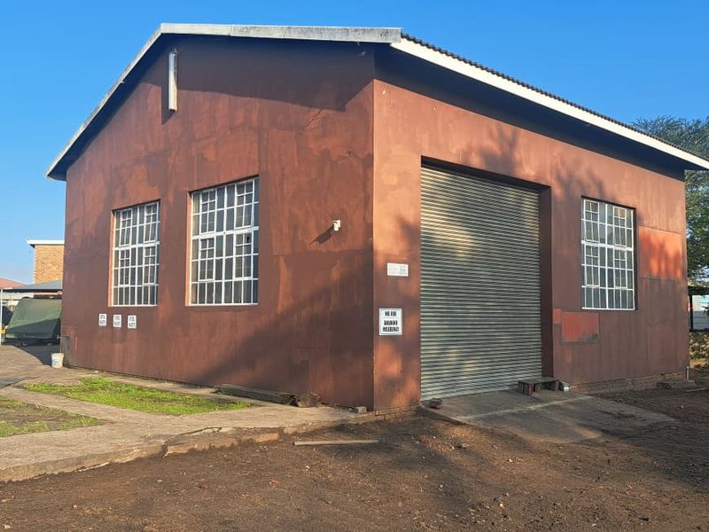 Industrial warehouse building featuring a large roller shutter door, steel-framed windows, and a durable metal roof, situated on a functional concrete and dirt lot.