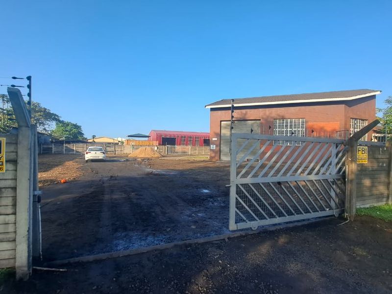 Industrial property entrance featuring a sliding gate, electric fencing, and a workshop building with a roller shutter door and unpaved yard space.