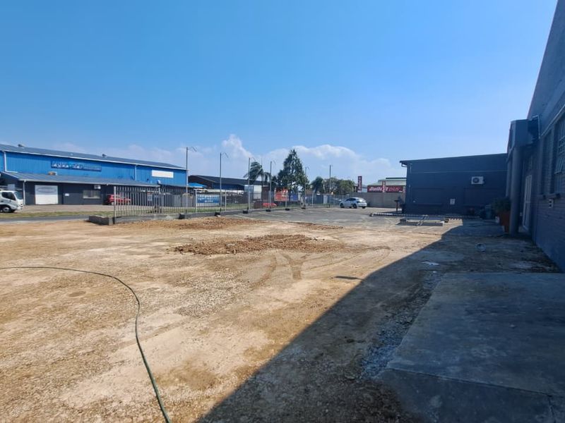 An expansive rear exterior area featuring a concrete apron, brick building facade, and metal security fencing, currently with an unpaved ground surface.