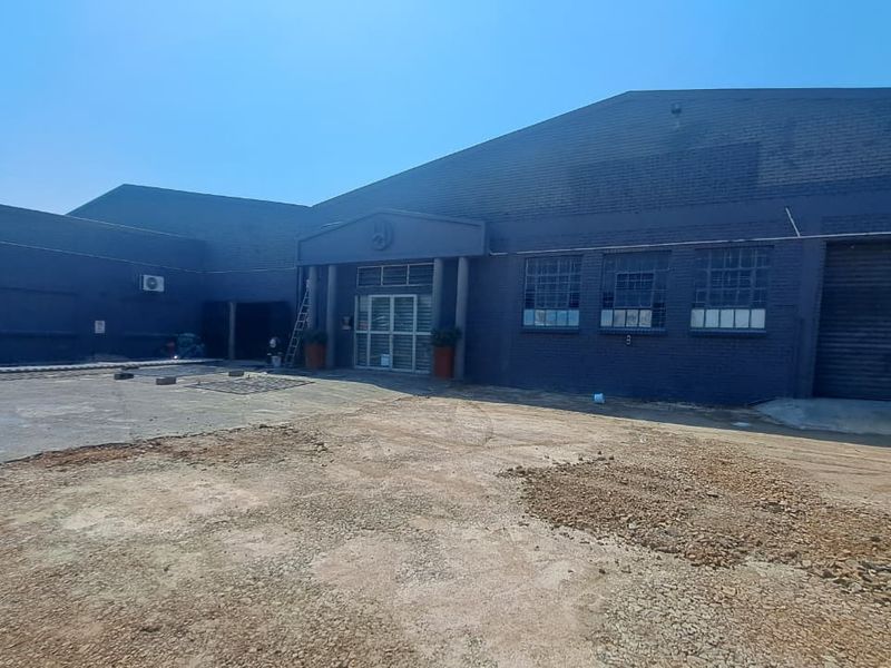 Front elevation of a commercial industrial property featuring a covered entrance, metal-framed windows, and a large roller shutter door with an unpaved forecourt.