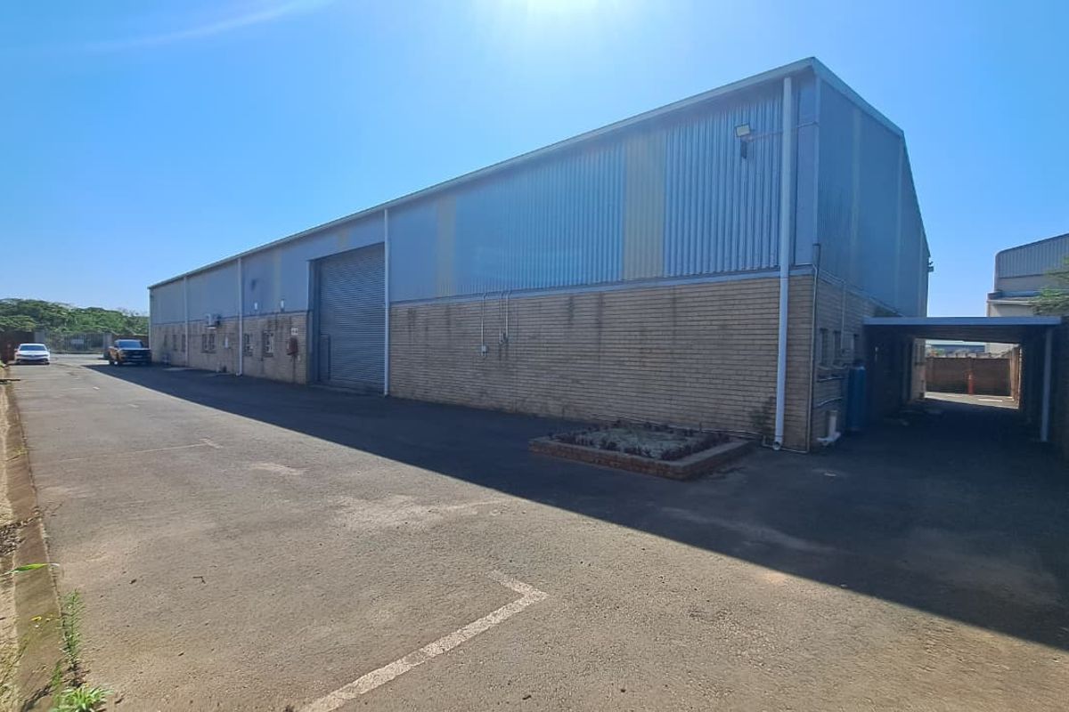 Exterior view of an industrial warehouse featuring corrugated metal cladding, brick facade, a large roller shutter door, and a paved driveway area.