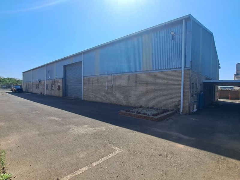 Exterior view of an industrial warehouse featuring corrugated metal cladding, brick facade, a large roller shutter door, and a paved driveway area.