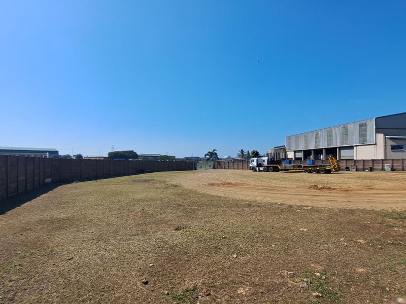 A wide-angle view of an industrial property featuring a large unpaved yard, enclosed by a precast concrete boundary wall with a warehouse structure visible.