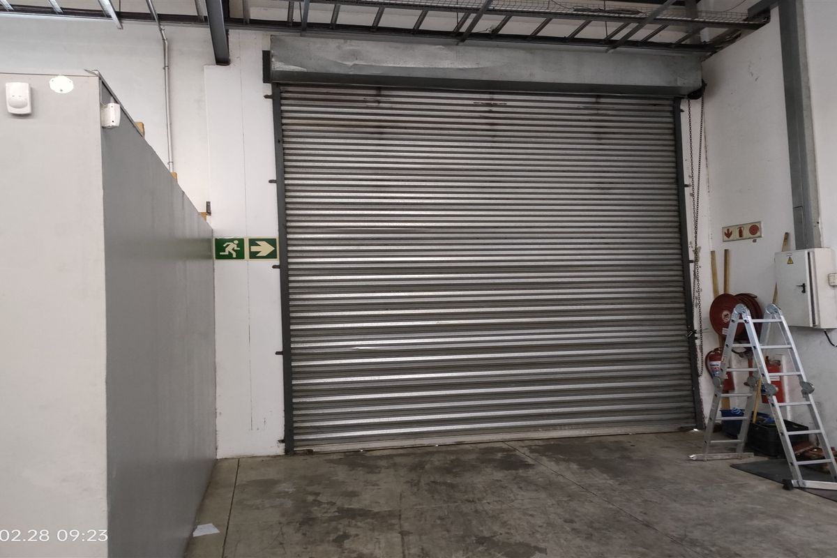 Industrial warehouse space featuring a large metal roller shutter door, exposed ceiling structure, and concrete flooring.