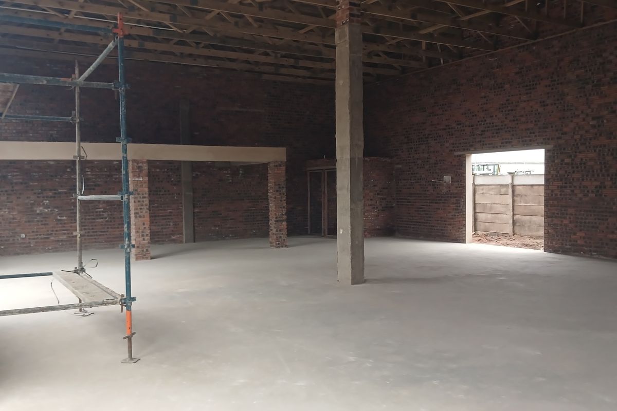 An expansive, unfinished industrial warehouse space featuring exposed brick walls, concrete flooring, and open timber roof trusses, currently under renovation.