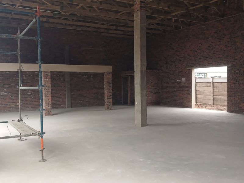 An expansive, unfinished industrial warehouse space featuring exposed brick walls, concrete flooring, and open timber roof trusses, currently under renovation.