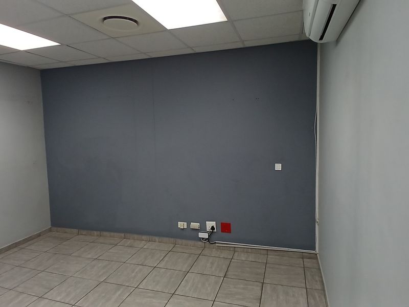 A clean, empty office space featuring tiled flooring, a suspended ceiling with recessed lighting, and a wall-mounted air conditioning unit.