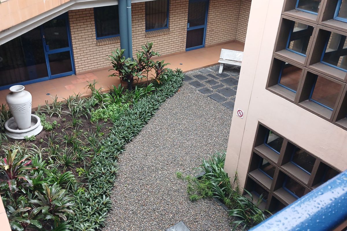 An elevated view of a landscaped industrial courtyard featuring a gravel path, garden beds, and brick building facades with blue-framed windows.