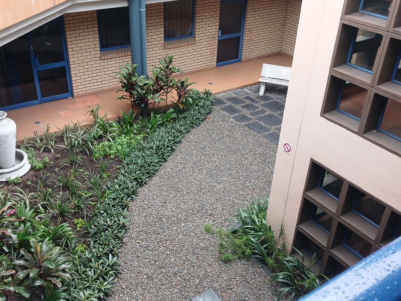 An elevated view of a landscaped industrial courtyard featuring a gravel path, garden beds, and brick building facades with blue-framed windows.