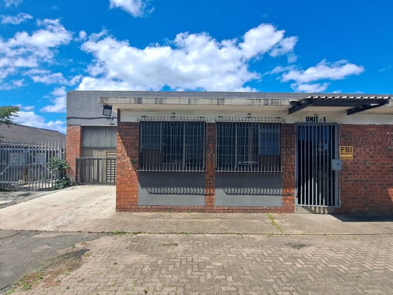 Front exterior view of an industrial unit featuring brick facade, security bars, metal gate, and paved driveway access.
