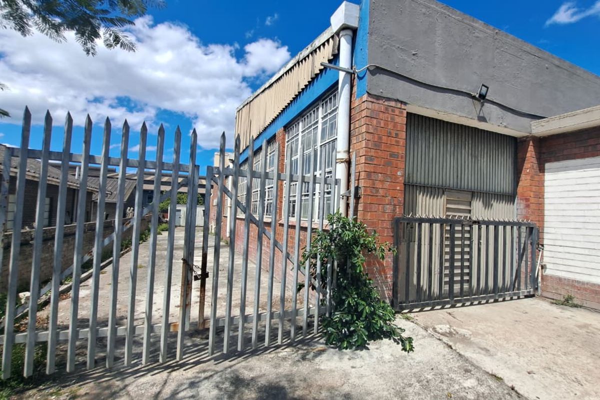 Rear exterior view of an industrial property featuring brick construction, steel-framed windows, a roller shutter door, and perimeter security fencing.