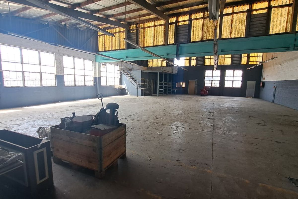 Spacious industrial warehouse featuring high ceilings with exposed steel trusses, a mezzanine level, concrete flooring, and large windows providing ample natural light.