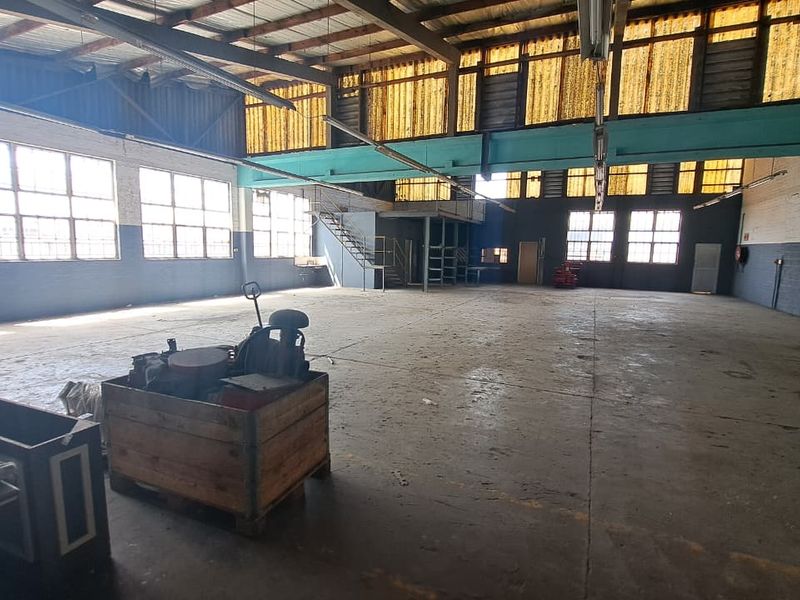 Spacious industrial warehouse featuring high ceilings with exposed steel trusses, a mezzanine level, concrete flooring, and large windows providing ample natural light.