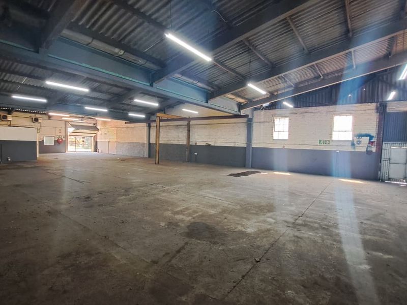 Spacious industrial warehouse unit featuring high ceilings with exposed steel trusses, concrete flooring, and large roller shutter access.