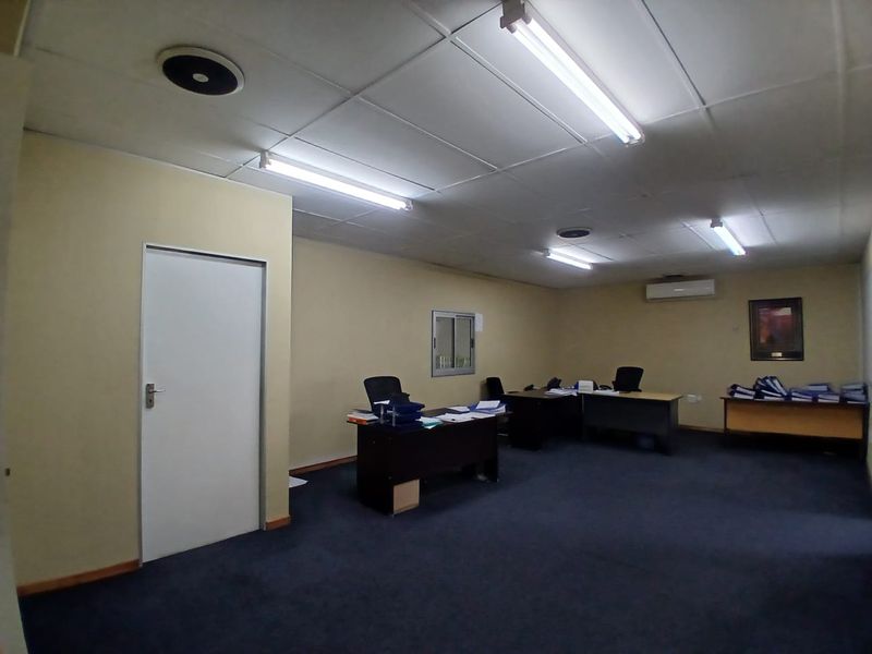 A functional office space featuring a suspended ceiling, fluorescent lighting, wall-mounted air conditioning, and carpeted flooring.