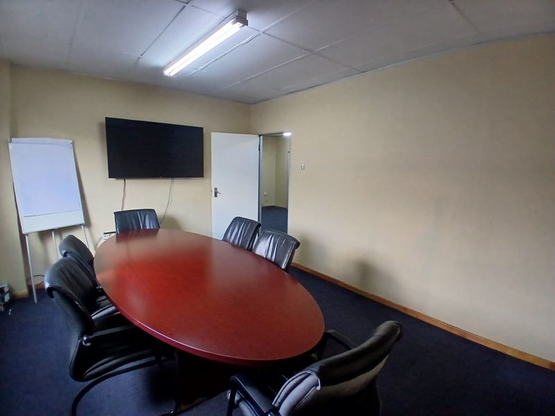 A functional office meeting room featuring carpeted flooring, a suspended ceiling with fluorescent lighting, and a central boardroom table setup.