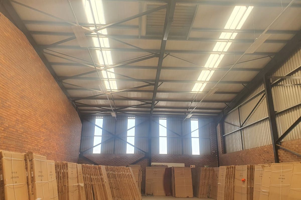 Spacious industrial warehouse featuring high ceilings with exposed steel trusses, brick walls, and large vertical windows providing ample natural light.